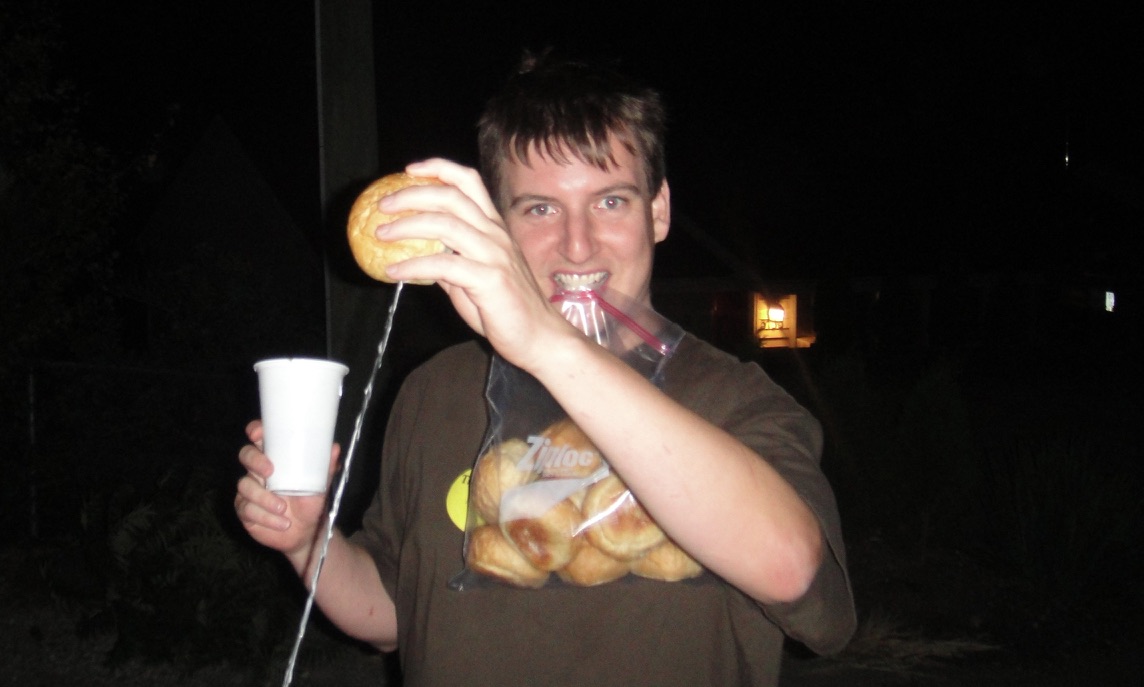 A smiling bunner with a roll, a cup, and a bag of buns at night.